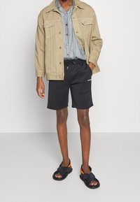 Chaqueta de mezclilla beige, camisa de rayas azul clara, pantalones cortos negros y sandalias oscuras con acentos naranjas. Diseño simple, corte relajado y colores básicos.