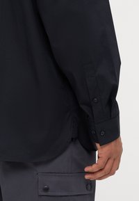 Camicia nera a maniche lunghe in tessuto liscio. Presenta un polsino con bottoni, orlo a coda di rondine e una sottile texture lucida. Abbinata a pantaloni cargo grigi.