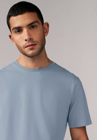 Světle modré tričko s krátkým rukávem, které má kulatý výstřih a hladkou texturu. Minimalistický design bez viditelných vzorů nebo akcentů.