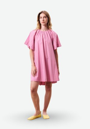 Jeune femme debout portant une robe rose à manches courtes amples avec de fines rayures et des chaussures plates jaunes sur un fond blanc.