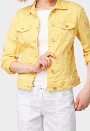 Vrouw die een felgele spijkerjack dichtknopen draagt over een wit shirt, met witte broek met trekkoord, tegen een effen achtergrond.