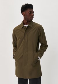 Olíva zöld vízálló kabát egyenes szabással, egyszerű gallérral, oldalsó zsebekkel és gombos záródással. Hossza ujjak és sima textúra jellemzi.