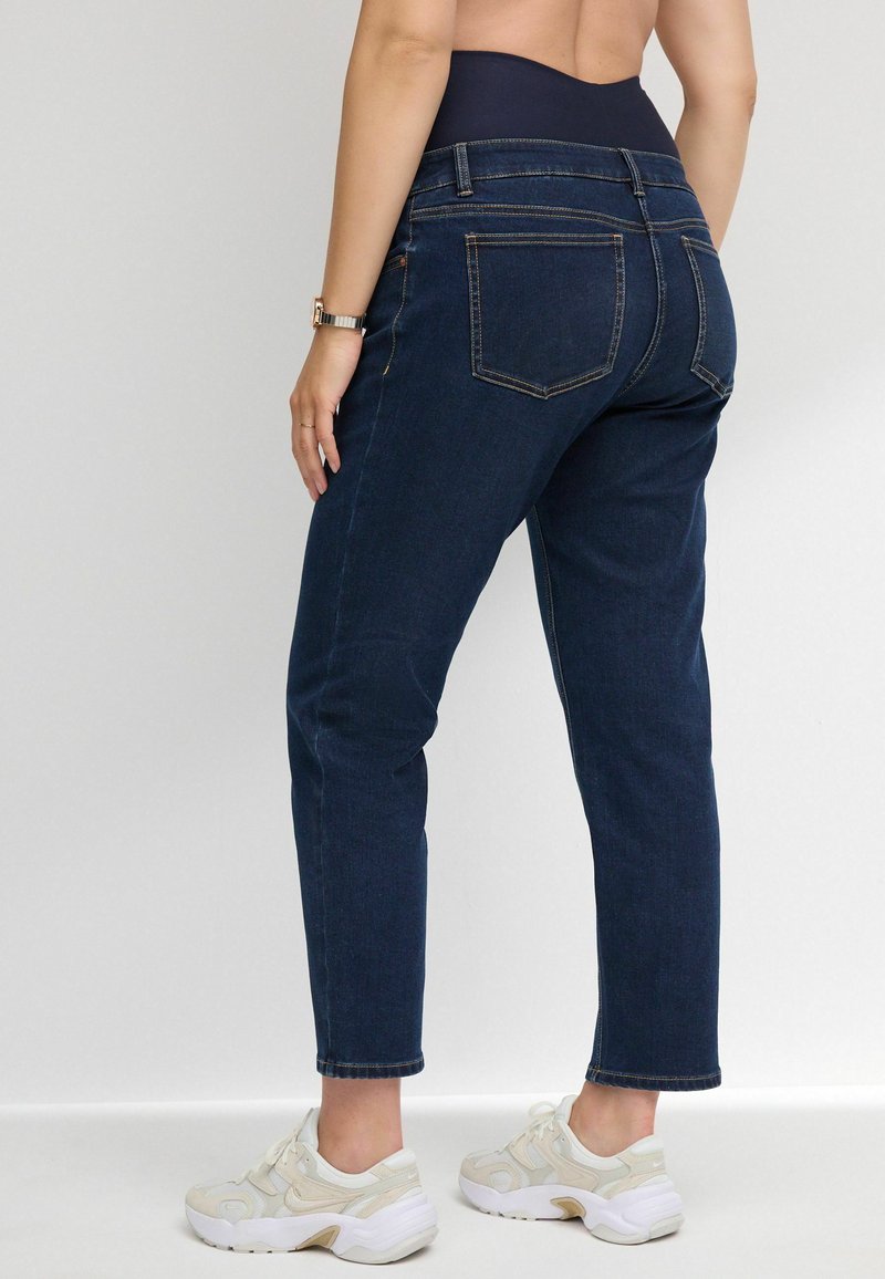 Jeans de mezclilla azul oscuro con pierna ajustada, cintura alta y dos bolsillos traseros. Lleva puestos unos zapatos deportivos gruesos de color claro.
