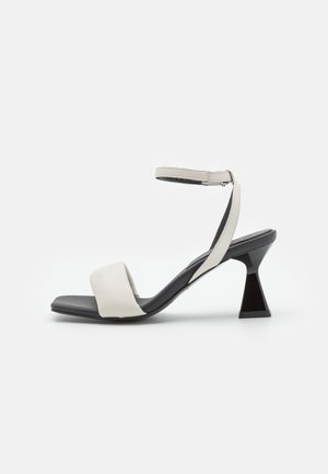 Beige Blockabsatzsandalen mit einem breiten Riemen über dem Zeh und dem Knöchel, ausgestattet mit einer eleganten schwarzen Sohle und einer geometrischen Absatzform.