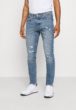 Hombre vistiendo jeans rotos de color azul claro, zapatillas blancas y una camiseta blanca, de pie frente a un fondo claro y liso.