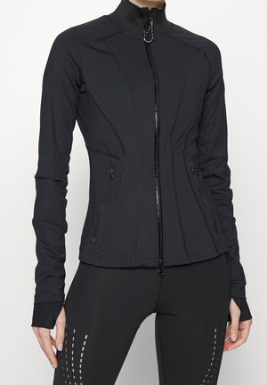 Vrouw draagt een nauwsluitende zwarte jas met ritssluiting en duimgaten, gecombineerd met bijpassende zwarte legging met verticale uitsnijdingspatronen.