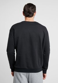 Man som vänder bort med en enkel svart sweatshirt med långa ärmar och ljusgråa sweatpants mot en neutral bakgrund.