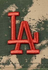 Green camo cap with a textured fabric featuring an embroidered red "LA" logo. Design includes scattered lighter pattern accents.