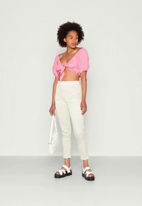 Top cropped rosa com mangas bufantes e laço na frente, combinado com calças justas brancas de xadrez e sandálias pretas de plataforma. Bolsa branca ao ombro.