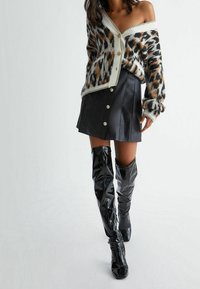 Cardigan à imprimé léopard avec boutons, associé à une mini jupe en cuir noir dotée de boutons à l'avant et des bottes noires en cuir brillant au genou.