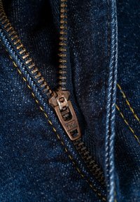 Nahaufnahme eines teilweise geschlossenen Reißverschlusses aus brünniertem Metall auf dunkelblauem Denim-Stoff mit gelben Nähten.