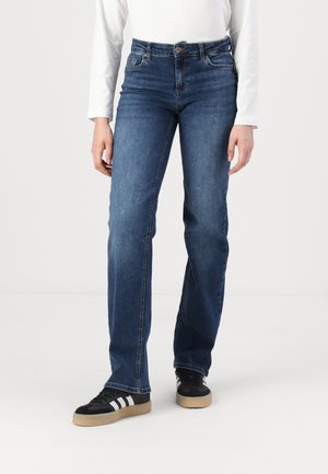 Dark blue jeans with a fitted design, made of denim, featuring a button closure and front pockets. Worn with black sneakers and white top.