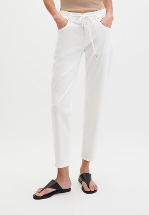 Person trägt weiße Kordelhose und schwarze offene Sandalen und steht mit den Händen in den Taschen vor weißem Hintergrund.