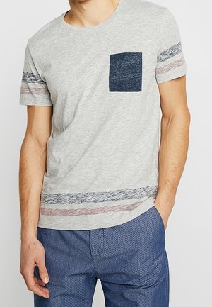 Mężczyzna noszący jasnoszary t-shirt z krótkim rękawem z ciemnoniebieską kieszenią na piersi i poziomymi paskami na rękawach i na dole, w połączeniu z niebieskimi spodniami.