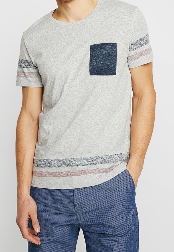 Homme portant un t-shirt gris clair à manches courtes avec une poche poitrine bleu foncé et des rayures horizontales sur les manches et l'ourlet, associé à un pantalon bleu.