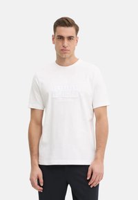 T-shirt bianco in cotone con maniche corte, caratterizzato da un logo testurizzato "TOMMY HILFIGER" sulla parte anteriore. Design semplice con scollo rotondo.