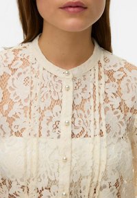 Koronkowa bluzka w kremowym kolorze z kwiatowymi wzorami, z zaokrąglonym kołnierzykiem i perłowymi guzikami z przodu. Przezroczysta faktura i delikatny wzór.