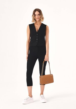 Femme aux cheveux ondulés mi-longs porte un gilet noir sans manches, un pantalon court noir, des baskets blanches et tient un sac à main marron avec des anses en chaîne.