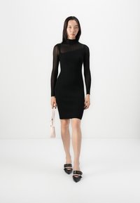 Black ribbed dress with long sleeves and high neckline. Model holds a light pink handbag, paired with black pointed-toe heels featuring straps.
