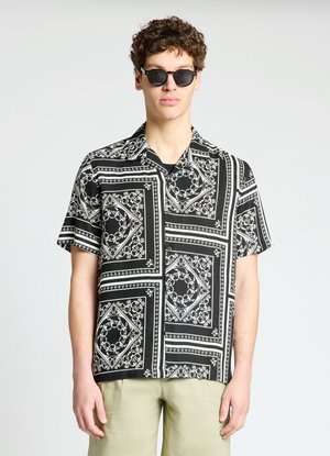 Jeune homme portant une chemise à manches courtes noire et blanche à motifs, un pantalon beige et des lunettes de soleil noires, debout devant un fond uni.