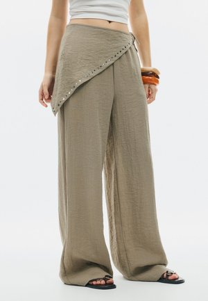 Osoba nosiaca voľné béžové nohavice s asymetrickým závinovým detailom a čierne sandále, doplnená tromi hrubými náramkami na zápästí.