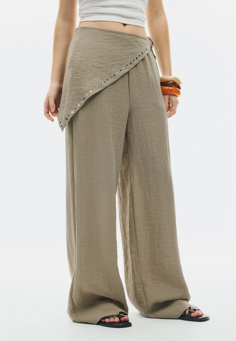 Personne portant un pantalon beige ample avec un détail d’enveloppe asymétrique et des sandales noires, accessoirisée de trois gros bracelets au poignet.