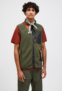 Fleece vest in olive green with a black nylon pocket, featuring two side zippers and an adjustable cord. Paired with a rust-colored shirt.