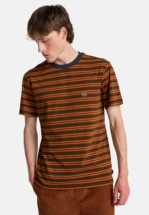 Jonge man met kort bruin haar, gekleed in een bruin-oranje gestreept T-shirt en een bruine broek, kijkend naar beneden links van hem.