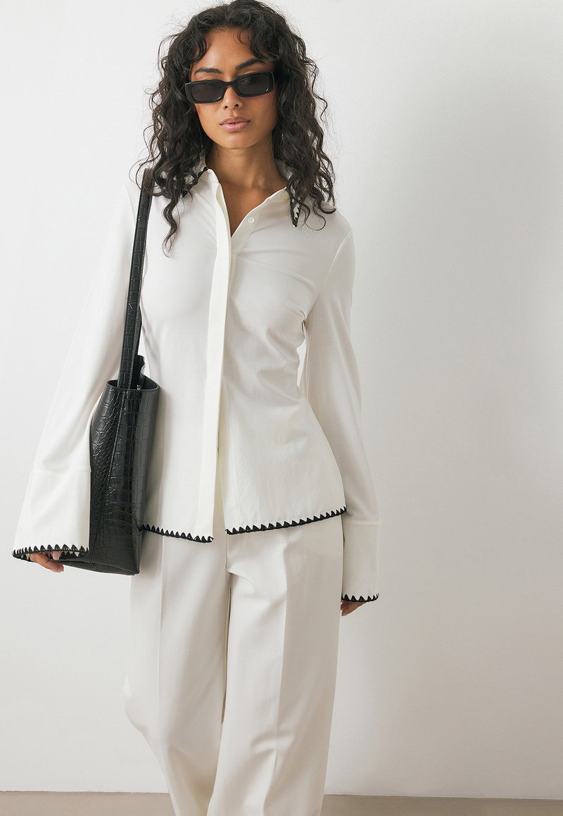 Weiße Bluse mit schwarzem gewellten Abschluss, langen Ärmeln, kombiniert mit weißen Hosen und einer schwarzen Ledertasche. Model trägt schwarze Sonnenbrille.