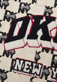 Écussons texturés DKNY et NEW en noir et blanc sur un fond en tissu beige avec des logos cœur NY noirs et gris répétés.