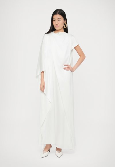 Bijela, lepršava maksi haljina s kratkim rukavima, asimetričnim naborima i glatkom teksturom. Uparena sa bijelim cipelama šiljatih vrhova.