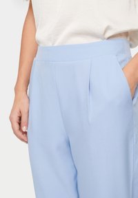 Světle modré kalhoty s vysokým pasem, s plisováním a bočními kapsami. Vyrobeno z hladkého materiálu s jemnou texturou.