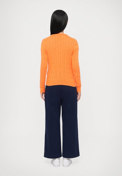 Oranžs džemperis ar kabeļu rakstu un gariem piedurkņiem, kopā ar platām tumši zilu biksēm. Apģērbu papildina vienkāršas baltas sporta kurpes.