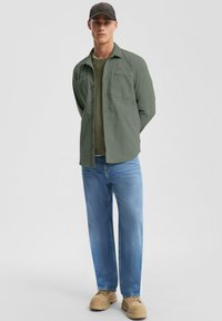 Lichtgroene overhemdd met twee voorzakken, gedragen over een groene t-shirt. In combinatie met blauwe jeans en beige casual schoenen.
