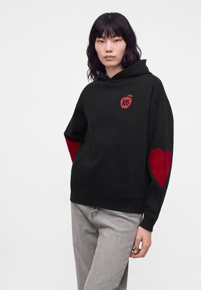 Jeune personne aux cheveux noirs portant un sweat à capuche noir avec un logo pomme rouge sur la poitrine et des patchs en forme de cœur rouges aux coudes, associé à un pantalon gris.