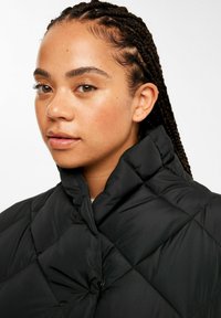 Schwarze gesteppte Pufferjacke mit hohem Kragen, glänzender Oberfläche und geometrischem Rautenmuster, die von einer Person getragen wird.