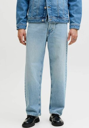 Mann trägt hellblaue Jeans mit geradem Bein, passende blaue Jeansjacke und schwarze Lederschuhe, steht entspannt mit Armen an den Seiten.