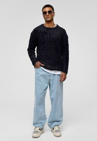 Marine kabelgebreide trui, relaxte pasvorm, ronde hals; lichtblauwe losse spijkerbroek; witte gelaagde t-shirt; witte sneakers; donkere zonnebril.