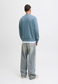 Férfi kék pulóvert, fehér aláöltözetet, laza világos koptatott farmert és fehér cipőt viselve, egy sima fehér háttér előtt állva, szemben a nézővel.