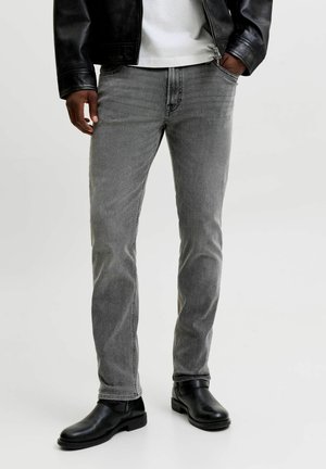 Homme portant un jean slim gris, des bottines Chelsea en cuir noir, une chemise blanche et une veste en cuir noire, les mains dans les poches sur fond blanc.