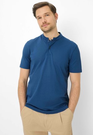Mann trägt ein blaues Henley-Shirt mit kurzen Ärmeln und beige Hosen, steht mit den Händen in den Taschen vor einem schlichten weißen Hintergrund.