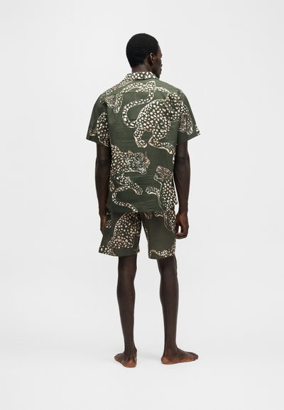 Άνδρας στέκεται ξυπόλητος, φοράει ταιριαστό πράσινο κοντομάνικο πουκάμισο και σορτς με μεγάλο φωτεινό στίγματα μοτίβο λεοπάρδαλης.