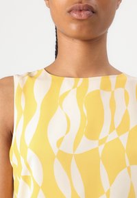Gros plan d'une personne portant un haut sans manches avec un motif abstrait ondulé jaune et blanc sur un fond uni.