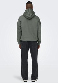 Sweat à capuche vert coupé avec une grande capuche, tissu texturé et poignets côtelés, associé à un jean en denim sombre ample et des baskets à semelle blanche.