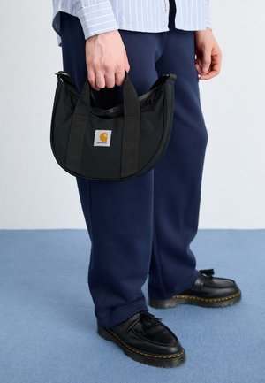 Person i marineblå bukser og sorte sko holder en lille sort Carhartt håndtaske med to håndtag og en lynlås på toppen.