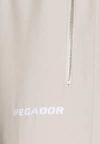 Béžová látka s hladkou texturou, vpravo se stříbrným zipem a vyšívaným logem "PEGADOR" v bílé barvě.