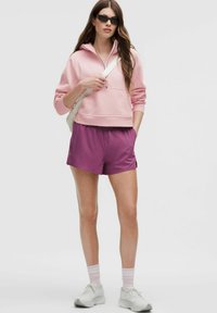 Helles rosa Zip-Up-Hoodie, lila Shorts, weiße Sneaker, rosa gestreifte Socken. Modell hält eine helle Tasche, Sonnenbrille, minimalistischem Design.