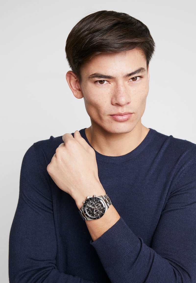 Montre chronographe en acier inoxydable avec un cadran noir, des maillons argentés et des boutons proéminents. Portée au poignet, main positionnée près du menton.