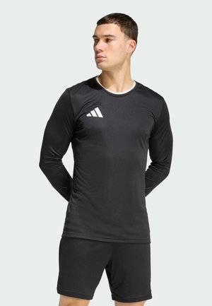 Mann trägt ein schwarzes langärmeliges Sportshirt mit weißem Logo und schwarze Shorts, steht mit den Händen hinter dem Rücken und schaut nach links.