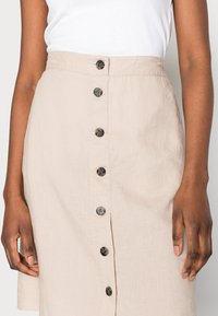 Jupe en lin beige avec une fermeture à boutons sur le devant, comportant cinq boutons foncés et une surface texturée. Main visible posée sur le tissu.